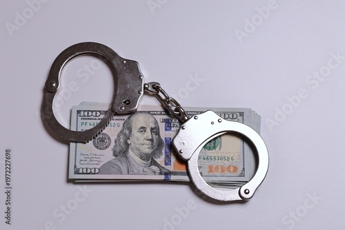 Metal handcuffs placed over a stack of US 100 dollar bills symbolizing financial crime, fraud, money laundering, tax evasion, and legal consequences. Strong conceptual image for topics like corruption