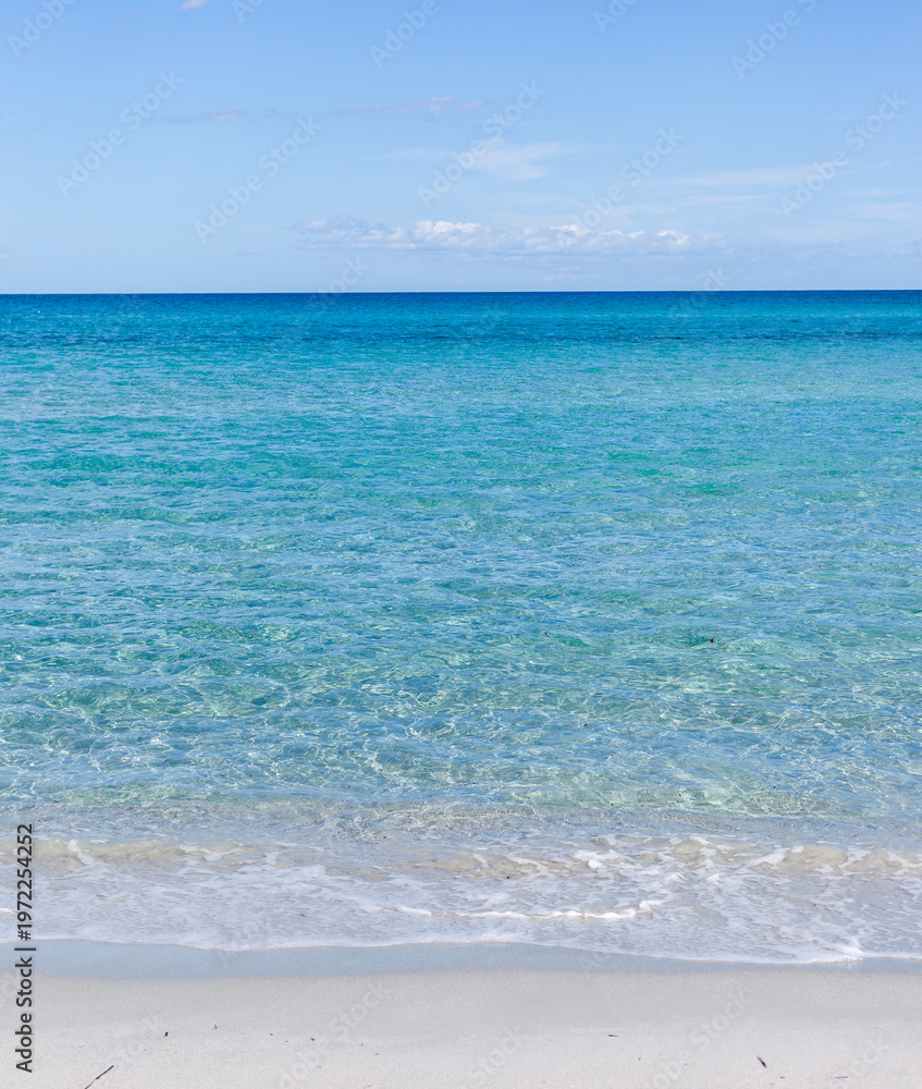 Fototapeta premium Turquoise sea waves gently lapping sandy beach shoreline in Sardinia, Italy. Calm Mediterranean waters under clear sky. Coastal summer landscape with pristine water.