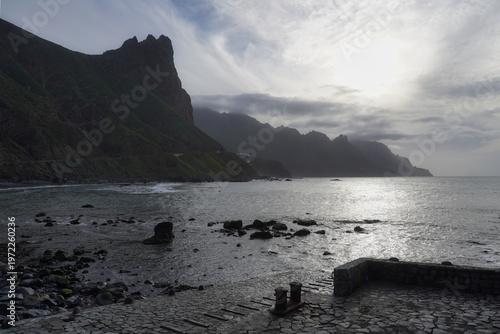 View of the dramatic coastal landscape of Anaga in Tenerife, with towering mountains, a volcanic stone pier and a bright silver sun reflection on the ocean surface under a misty sky