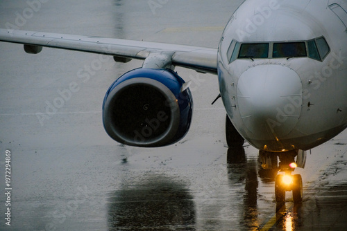 Striking front viewpoint focusing on the smooth aerodynamic nose and massive wing engine of a commercial jet resting on the highly reflective soaked airport apron
