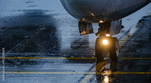 Close up view of an aircraft front wheels and bright landing lights shining upon a soaked concrete runway surface displaying strong reflections in damp weather