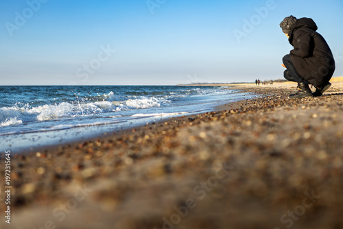 Foto Woman in winter clothing on beach searching for stones on shoreline, leaning for