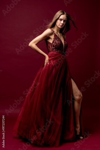 Model in long burgundy dress poses confidently in studio with dark red background showcasing the garment's design