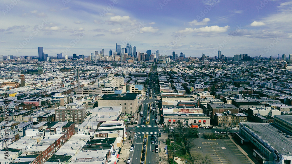 Obraz premium Central corridor ascent along Broad St reveals dense South Philly blocks converging toward the Center City skyline. Reflective roofs, brick gradients, soft atmospheric haze settle over Philly, PA