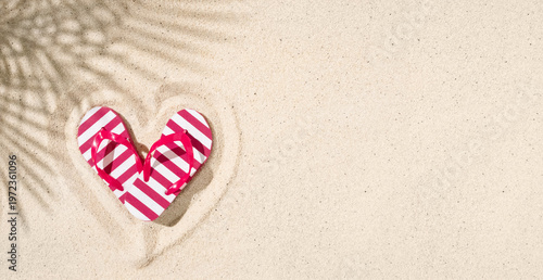 Colorful striped flip flops arranged in heart shape on sandy beach with palm shadow