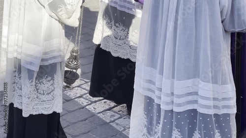 Young altar servers acolytes dressed in white lace robes and black vestments standing in sunlit outdoor religious ceremony traditional Christian Catholic procession setting