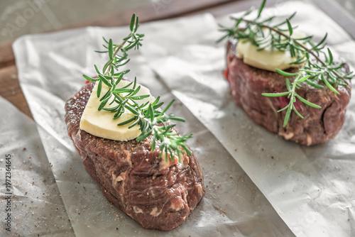 Two raw beef steaks with rosemary and butter on parchment paper over wooden board rustic kitchen
