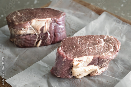 Two raw beef steaks on parchment paper over wooden board in natural light rustic kitchen setting