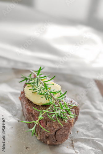 Single raw beef steak with rosemary and butter close up on parchment paper rustic kitchen