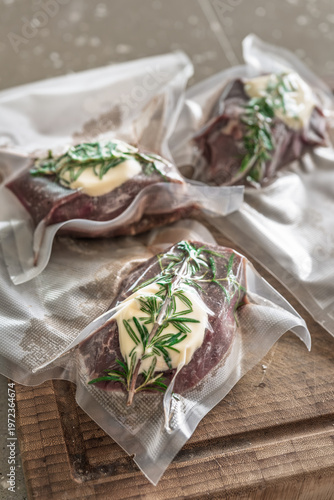 Three raw beef steaks with rosemary and butter in vacuum bags on wooden board sous vide