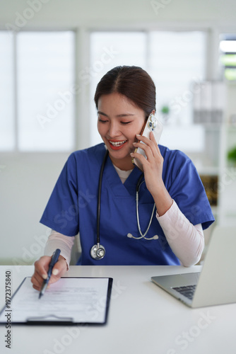 Asian adult female doctor nurse healthcare professional smiles uses smartphone call writes medical document works office clinic environment