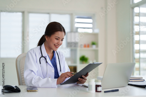 Schilderij op canvas Asian adult female doctor smiles reviewing patient medical records on clipboard