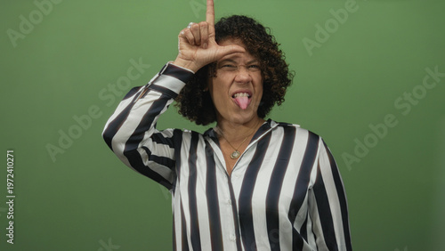 Fotografi Woman smirking while fingers form l gesture on forehead in green studio wearing striped blouse; playful mockery