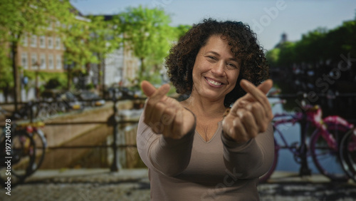 Schilderij op canvas Hispanic woman with curly hair and smiling expression makes finger heart gesture with hands on street near canal bridge; affection connection warmth joy