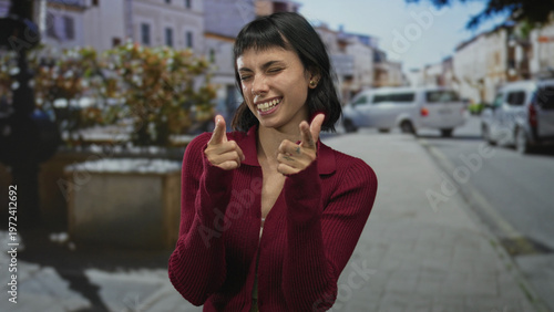 Photography Young hispanic woman with smiling face pointing fingers at camera on urban street; playful confidence