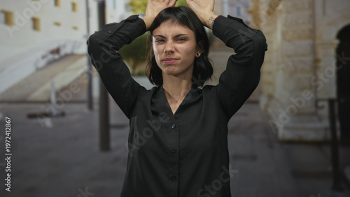 Fotografia Young hispanic woman making rabbit ears gesture with bare hands atop head on bustling street; playfulness