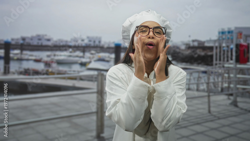 Εκτύπωση καμβά Woman chef cupping hands at open mouth shouting on a building terrace by a marina wearing chef hat and glasses; excitement celebration