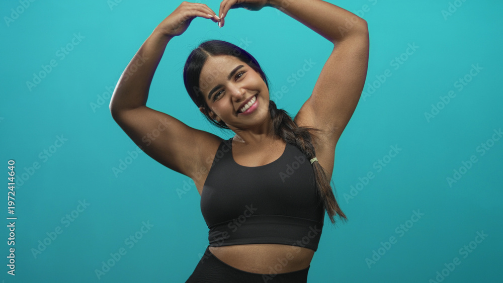 Fototapeta premium Young hispanic woman with bare arms and hands forms heart with fingers above her head while smiling, wearing a black sports bra in a studio setting; joy wellness positivity.