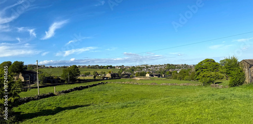 Expansive green fields stretch across rolling hills with dotted houses in the background. The clear blue sky enhances the peaceful rural setting near Thornton, Yorkshire, UK