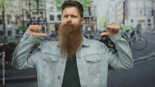 Man with long beard wearing denim jacket pointing thumbs to chest amidst blurred city street scene; pride.