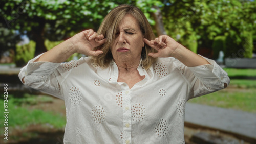 Billede på lærred Woman in white eyelet blouse covers her ears with fingers while grimacing among trees in forest; annoyance irritation distress upset