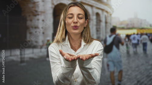 Photography Woman blowing a kiss with cupped hands in front of the colosseum building in rome among tourists; playful romance