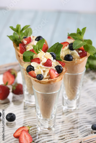 Appetizing dessert made of thin crepes rolled into cones  placed in shot glasses, filled with whipped cream, fresh strawberries, blueberries, almond flakes, and mint leaves