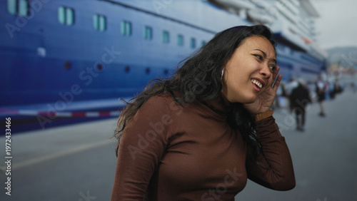 Konstfotografi Young hispanic woman holding hand to ear beside large blue cruise ship at port on street, looking over shoulder in alarm; concern travel delay