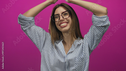 Принти на полотні Woman wearing striped shirt and glasses raises arms overhead in studio with vibrant pink background and broad smile; joy excitement