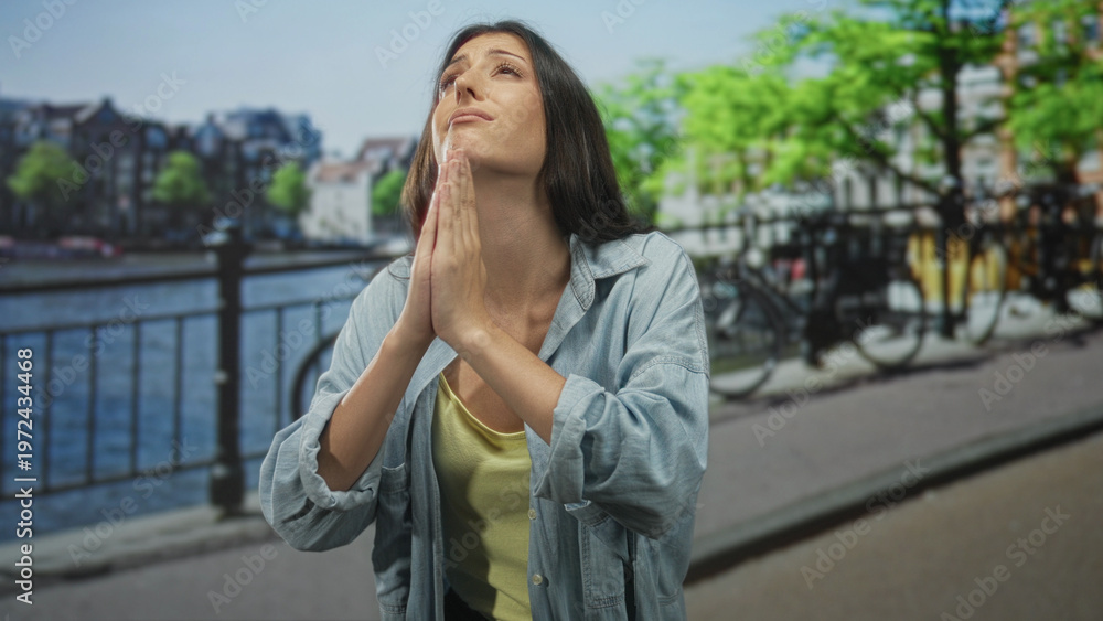 Fototapeta premium Young hispanic woman with hands clasped in prayer and closed eyes on amsterdam street by canal railing, wearing denim shirt; hope plea.