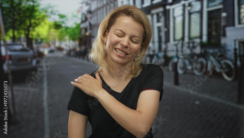 Tableau sur toile Blonde woman holds left shoulder on busy cobblestone city street lined with parked cars and bicycles under sunlight; discomfort