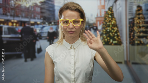 Obraz na plátně Young blonde woman waves hand while standing on busy city street during holiday season; greeting warmth