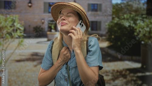 Fotografi Woman wearing pith helmet holding smartphone to ear at sunlit building entrance outdoors; adventure exploration wanderlust relief