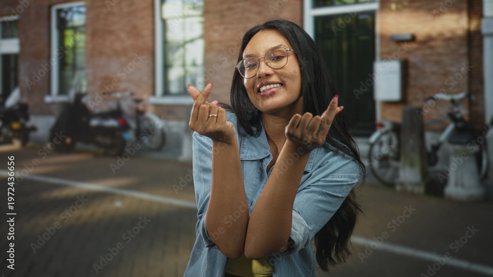 Fototapeta premium Woman points fingers at cobblestone street, makes finger heart and beckoning gestures while smiling, wearing glasses and denim shirt; playful charm.