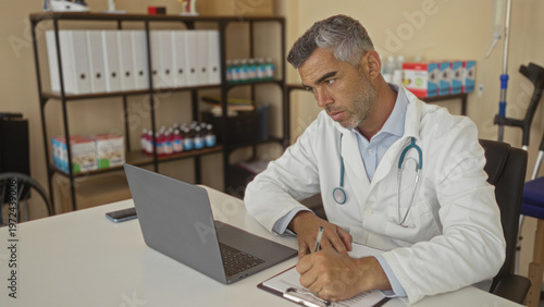 Foto Man doctor in white coat writing on clipboard at desk in medical building with laptop and stethoscope, crutches and medicine shelves behind him; focused care