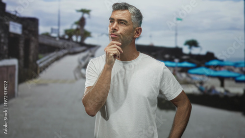 Quadro em tela Man hand on chin pensive pose on beach promenade with blue umbrellas and walkway; thoughtful reflection