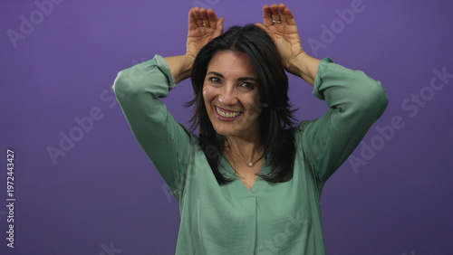 Obraz na plátně Hispanic woman wearing a green blouse smiling with hands raised behind head against a purple studio wall; playfulness