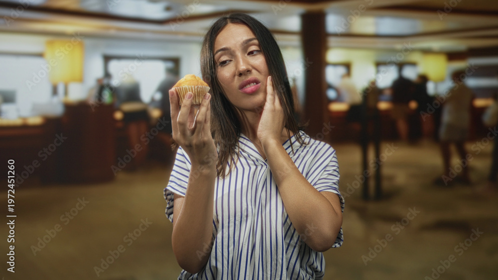 Fototapeta premium Young hispanic brunette woman holding muffin, hand on cheek, wearing striped shirt in hotel building; toothache discomfort.