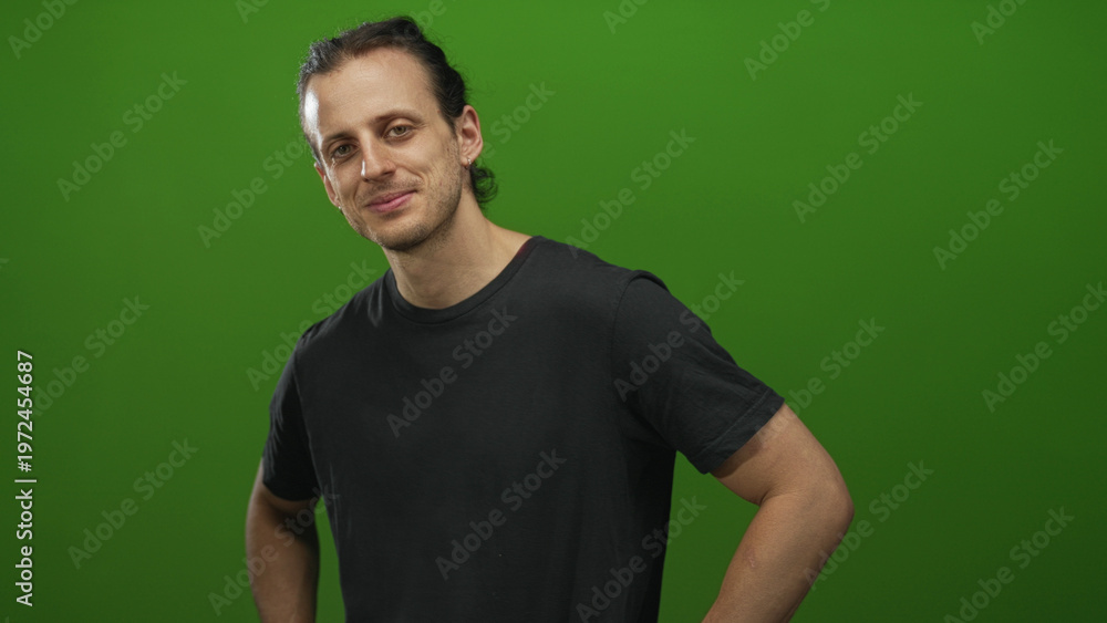 Fototapeta premium Man with hands on hips and visible bare forearms in studio with green backdrop, slight smile and relaxed posture; confident approachable.