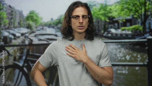Man, young hispanic with long hair and hand to chest on street by amsterdam canal, wearing glasses and gray t shirt, looking pensive; sincerity introspection.