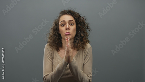 Photos Young hispanic woman pressing hands together in pleading gesture wearing beige top in gray studio; remorse apology