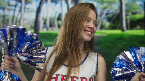 Photography Young caucasian woman shakes blue and silver pom poms energetically while smiling in green park; team spirit joy