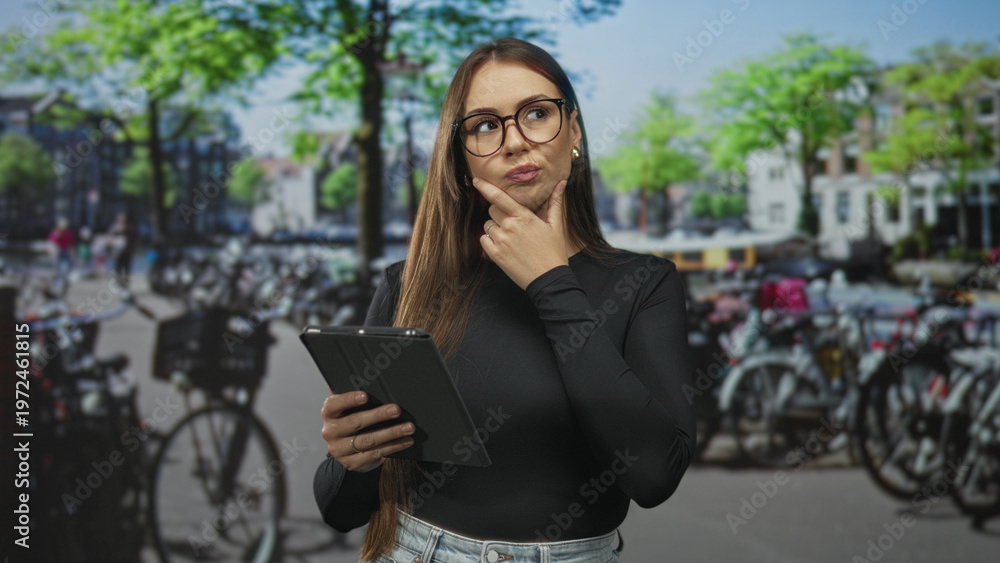 Fototapeta premium Woman wearing glasses and black top taps tablet with finger while holding device against a studio backdrop of parked bicycles; thoughtful.