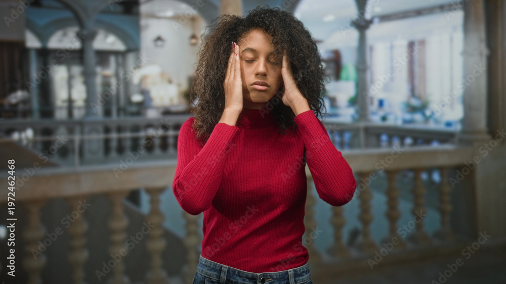Fototapeta premium Teen woman with curly hair pressing hands to temples in ornate building wearing red sweater; stress headache.
