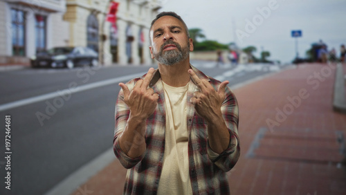 Man shows middle fingers to camera on street wearing plaid shirt and beard visible; urban defiance attitude.