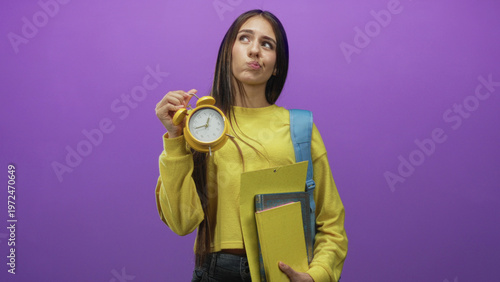 Cuadro en lienzo Teenage student holds yellow alarm clock and notebooks, pouts while carrying backpack in studio; time pressure frustration
