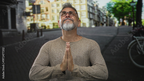 Tablou pe pânză Man with palms pressed together praying on a street, glasses and beard visible, hands clasped in front; penitence hope
