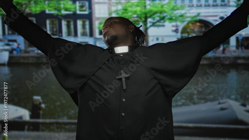 Εκτύπωση καμβά Man priest wearing clerical collar and cross necklace, arms outstretched on a street by a canal; devotion reflection serenity