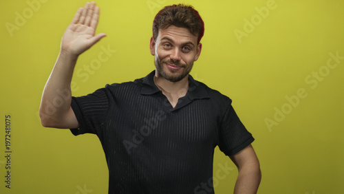 Foto Man raising right palm clearly visible against bright yellow studio wall, wearing black polo shirt with casual stance and slight smile; friendly greeting