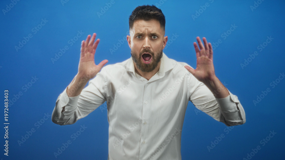 Fototapeta premium Man with beard and white shirt, hands raised and open mouth showing teeth, gesturing palms toward camera in studio; frustration.